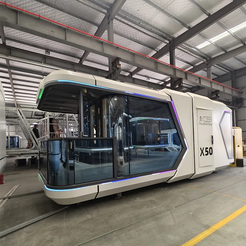 A futuristic white modular pod or cabin with large tinted glass windows and blue LED lighting accents, photographed inside a large industrial warehouse or factory. The side of the unit shows the text 'X50' and 'AERONAUTICS'.