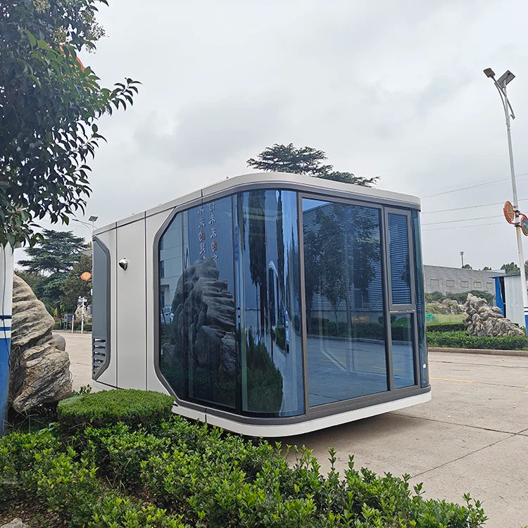 A modern small modular office or guardhouse with a white exterior and large dark-tinted glass windows, photographed outdoors on a paved area with shrubs, trees, and a cloudy sky in the background.