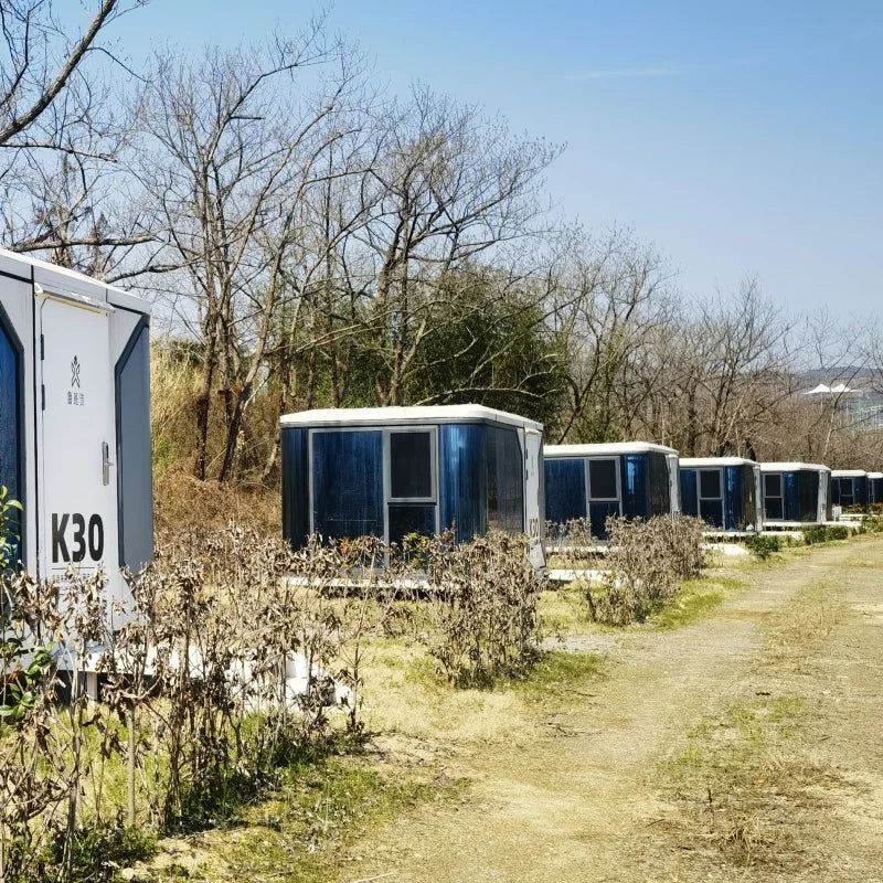 A row of modular portable toilets in blue and white containers parked outdoors on a dirt path with dry grass and leafless trees in the background.