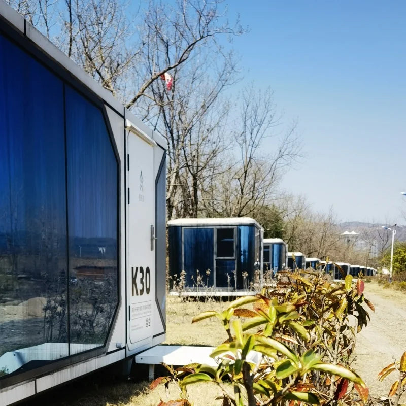 A row of modular portable toilets in an outdoor setting with trees and a dirt ground under a clear blue sky.
