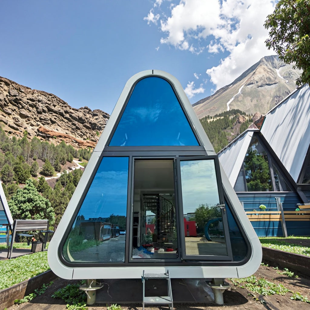 A modern triangular A-frame prefab cabin with large reflective blue-tinted windows and a central glass door, photographed outdoors in front of mountains and other cabins under a blue sky.