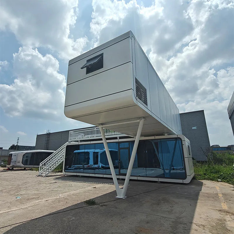 A modern elevated modular container home with a white exterior, large glass windows, and angled support legs, photographed outdoors on a concrete lot under a cloudy sky.